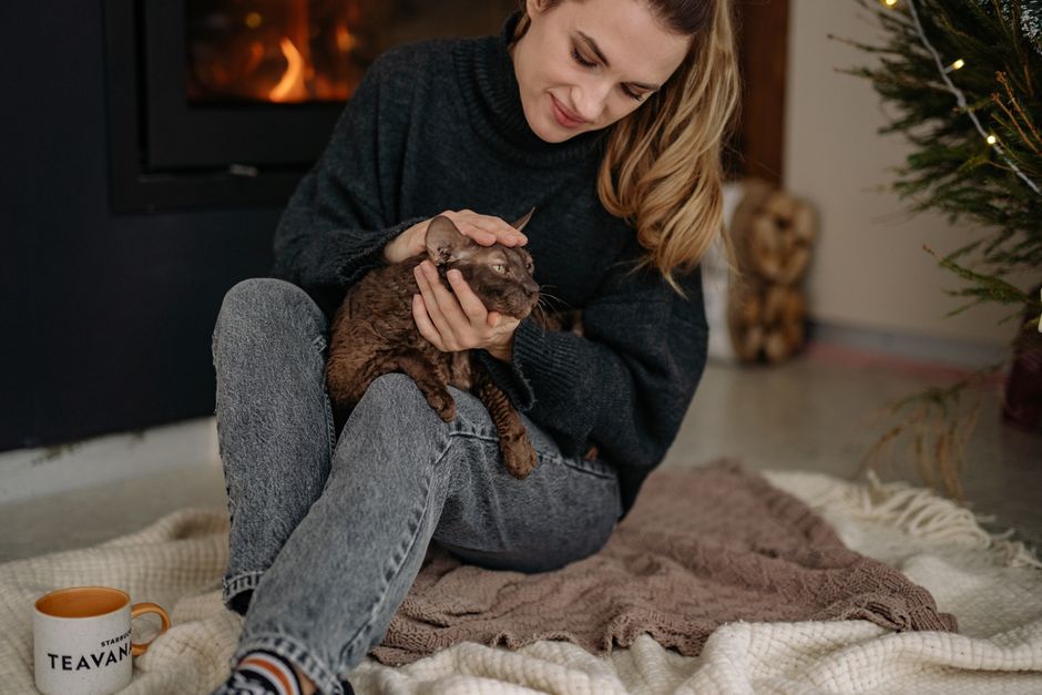 Woman in a sweater sitting on a rug by a fireplace, petting her cat